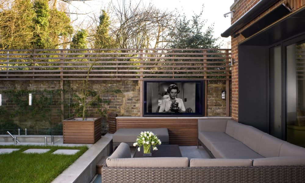 <span class="uk-h4">A backyard patio with an outdoor TV built into a wall structure with plants and trees behind it.</span>